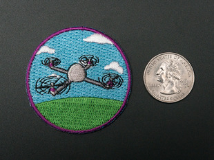 Circular embroidered badge showing a small grey drone over a stylized blue sky, with white clouds, and green grass. Badge is trimmed in purple and shown next to a quarter for scale. 