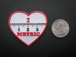 Heart shaped embroidered badge with the numbers 1, 2, 3 and the markings of a ruler in black over a white background. The words I and METRIC in red above and below the ruler. The badge is trimmed in red and shown next to a quarter for scale. 