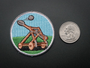 Circular embroidered badge with brown catapult mid-fling over an abstracted blue sky and green grass background. Badge is trimmed in grey and shown next to a quarter for scale. 