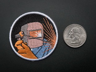 Circular embroidered badge showing the bust of an active welder in grey mask, orange safety gloves and blue coveralls with a corona of radiating orange sparks, over a black background. Badge is trimmed in white and shown next to a quarter for scale. 