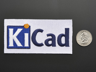 Rectangular embroidered badge with the letters KICA in blue and black on a white background, next to a quarter for scale. 