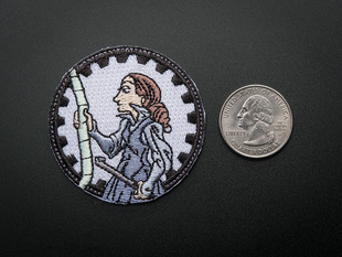 Circular embroidered badge showing a cartoon of Ada Lovelace in profile, holding a crowbar and examining a long ruler, over a white background. Badge is edged in black gear teeth. Shown next to a quarter for scale. 