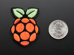 Embroidered patch in the shape of the raspberry pi logo, in red with green leaves on top, outlined in black. Shown next to quarter for scale. 