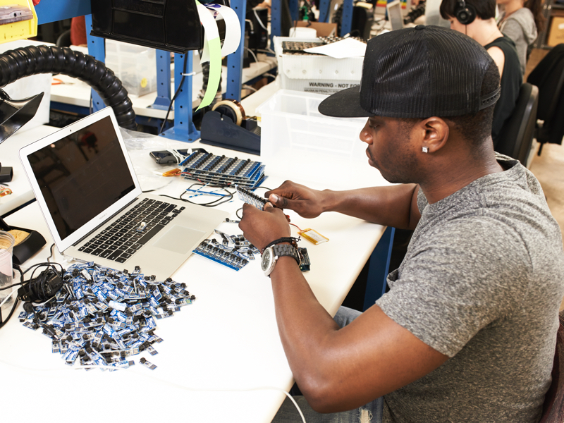 Young man with baseball cap, at electronic work bench, breaking apart a grid of small electronic boards