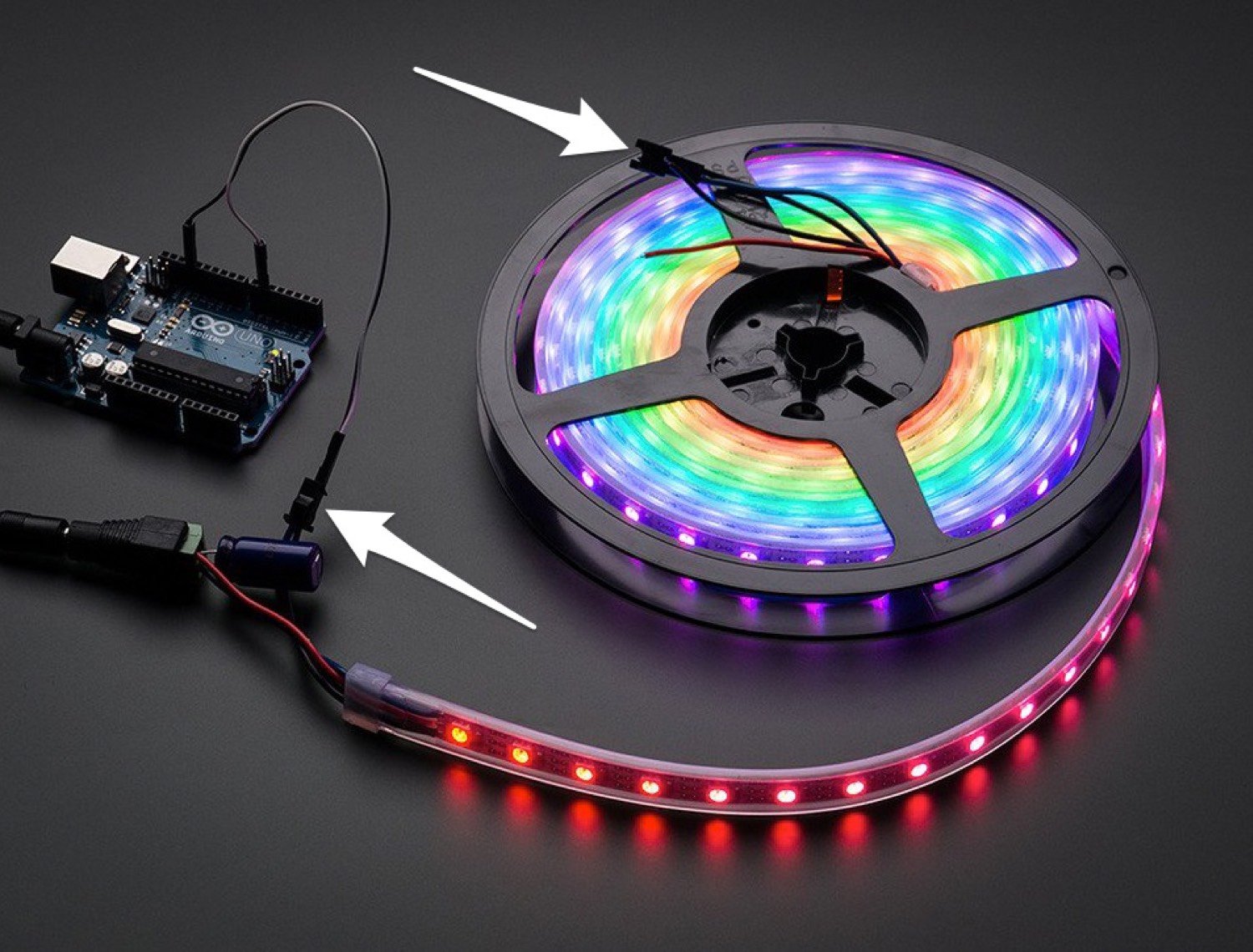 Full-length RGB LED strip in a spool, lit up in rainbow colors. Diagram arrows indicate the LED strip's built-in connectors. One at the end of the spool connects to an Arduino which is driving the strip. The other connector is at the center of the spool.
