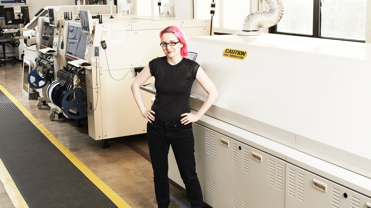 Woman with pink hair standing in front of electronic manufacturing equipment, arms akimbo.